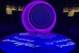 The setting for the Youth Theatre show back 2024 in the New Vic auditorium. It is lit in purple and features an eclipse backdrop and words projected on the floor including 'and they all lived happily ever after?', 'The Young Good Wolf' and 'The Trickster's Downfall'