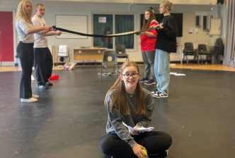 Youth Theatre members in rehearsal for the 2024 show back. One performer sits in the foreground cross-legged with a script and holding a tennis ball while four other performers behind her hold pieces of fabric between them, creating a bridge-like structure.