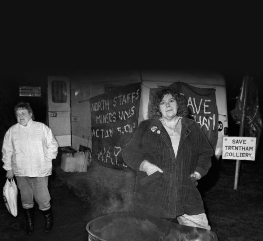 A black and white photograph of two woman in front of a caravan with various signs and banners with phrases such as 'North Staffs Miners Wives Action Group' and 'Save Trentham Colliery'. Credit Kevin Hayes