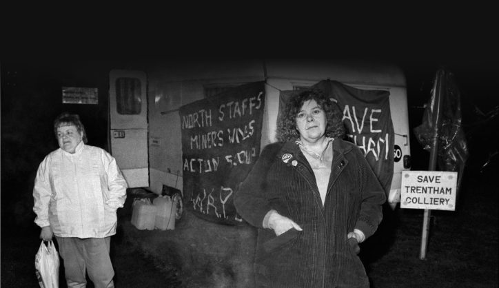 A black and white photograph of two woman in front of a caravan with various signs and banners with phrases such as 'North Staffs Miners Wives Action Group' and 'Save Trentham Colliery'. Credit Kevin Hayes