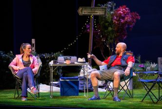 A heterosexual couple sit on some camping chairs having a discussion over a foldable table, surrounded by camping equipment, foliage, fairy lights and a footpath sign.