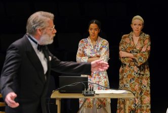 A man in a tuxedo gestures with a hand as two unimpressed women in dressing gowns with a laptop and script on a desk in front of them look at him.