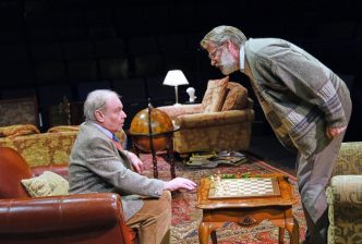 An older man sitting in a leather armchair looks into the distance with a sense of shock as another man sits up from his chair and talks to him, as if checking he's okay