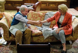 A man dressed smart-casually with an apron over the top, and a woman wearing a blazer, sit on a floral sofa and shake hands