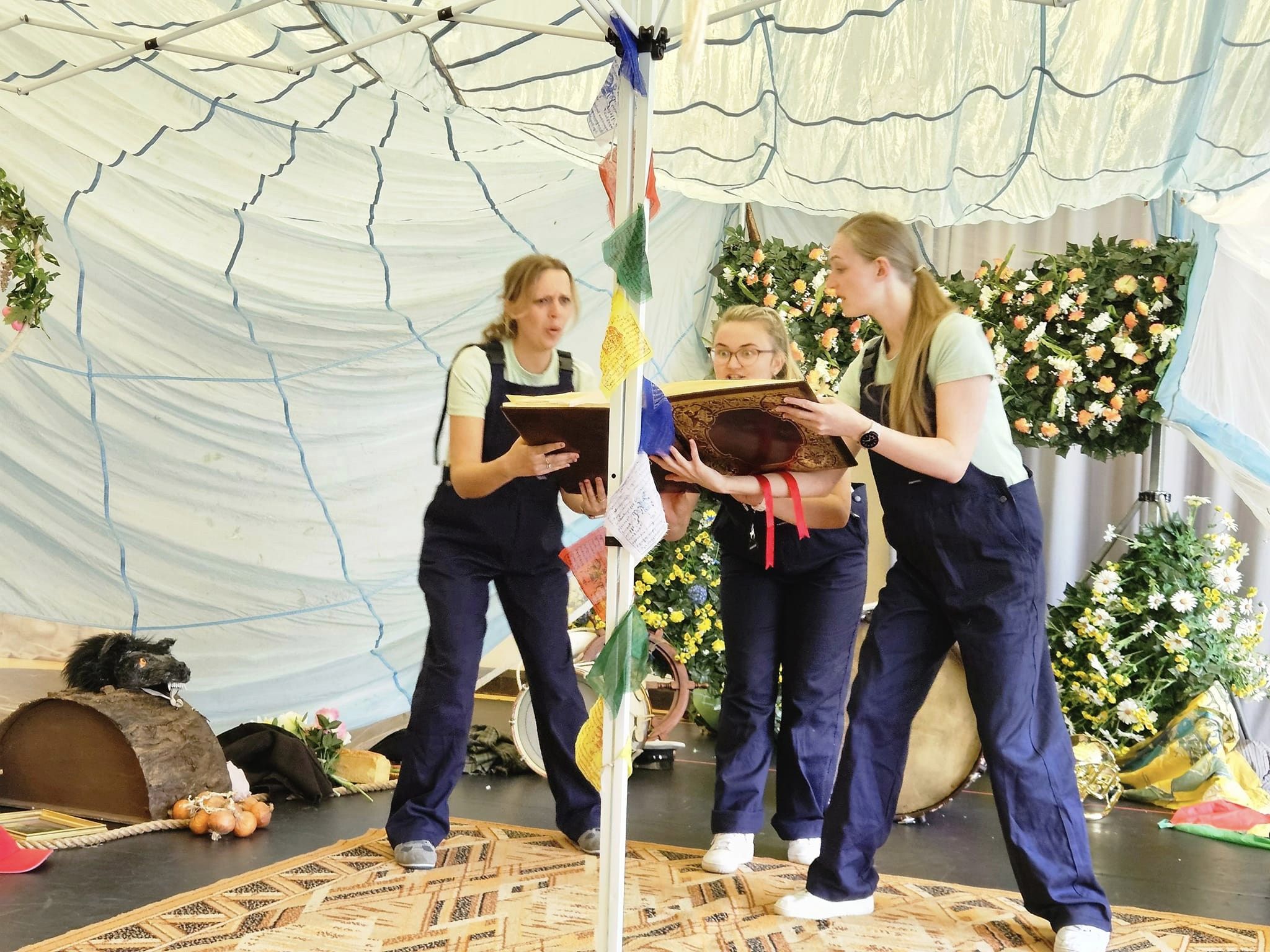 Three performers dressed in dungarees tell a fairy tale story holding a big book. They are surrounded by various props and foliage inside a tent.